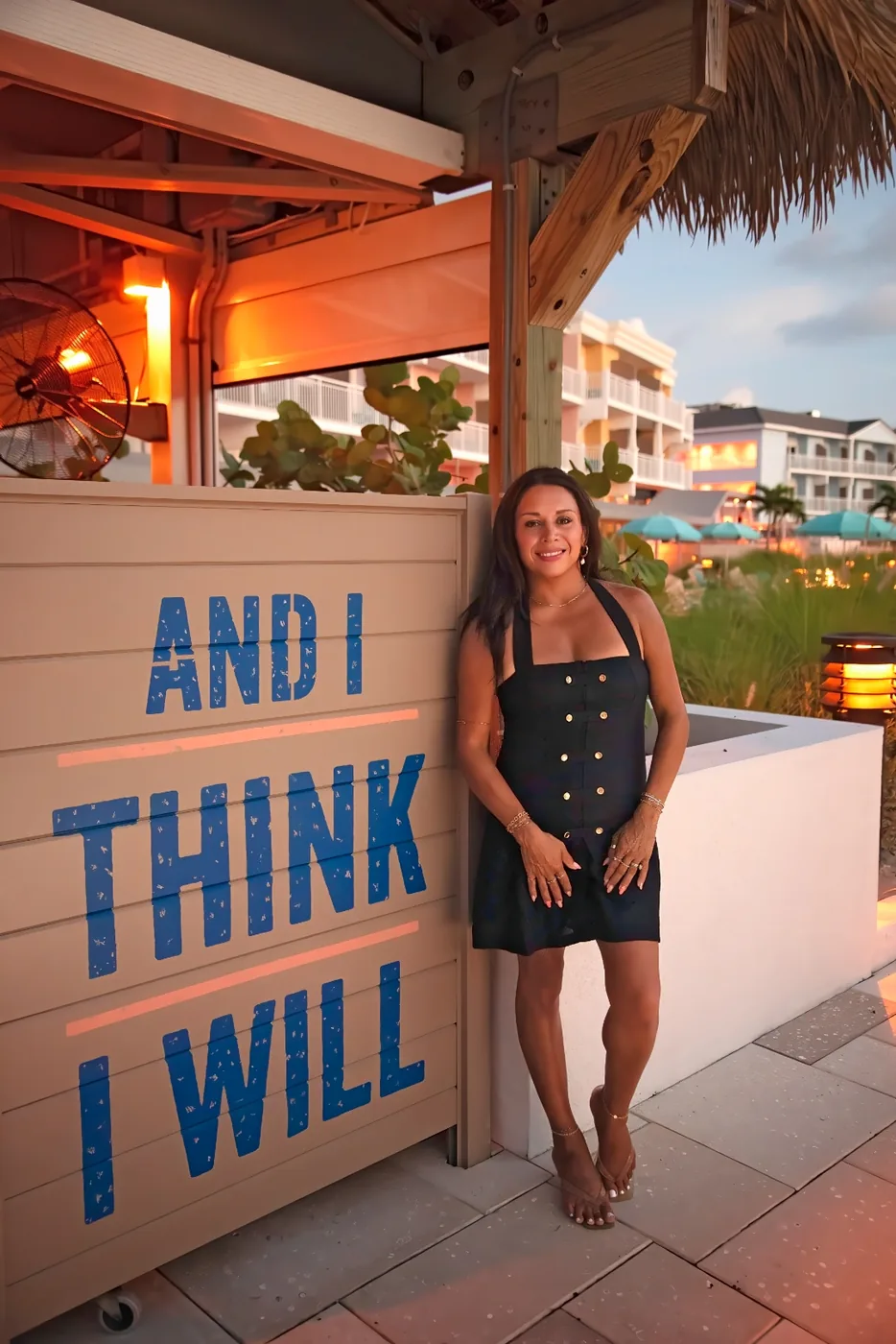 Emmy Paola Akin beside an And I Think I Will wall sign at the resort at dusk