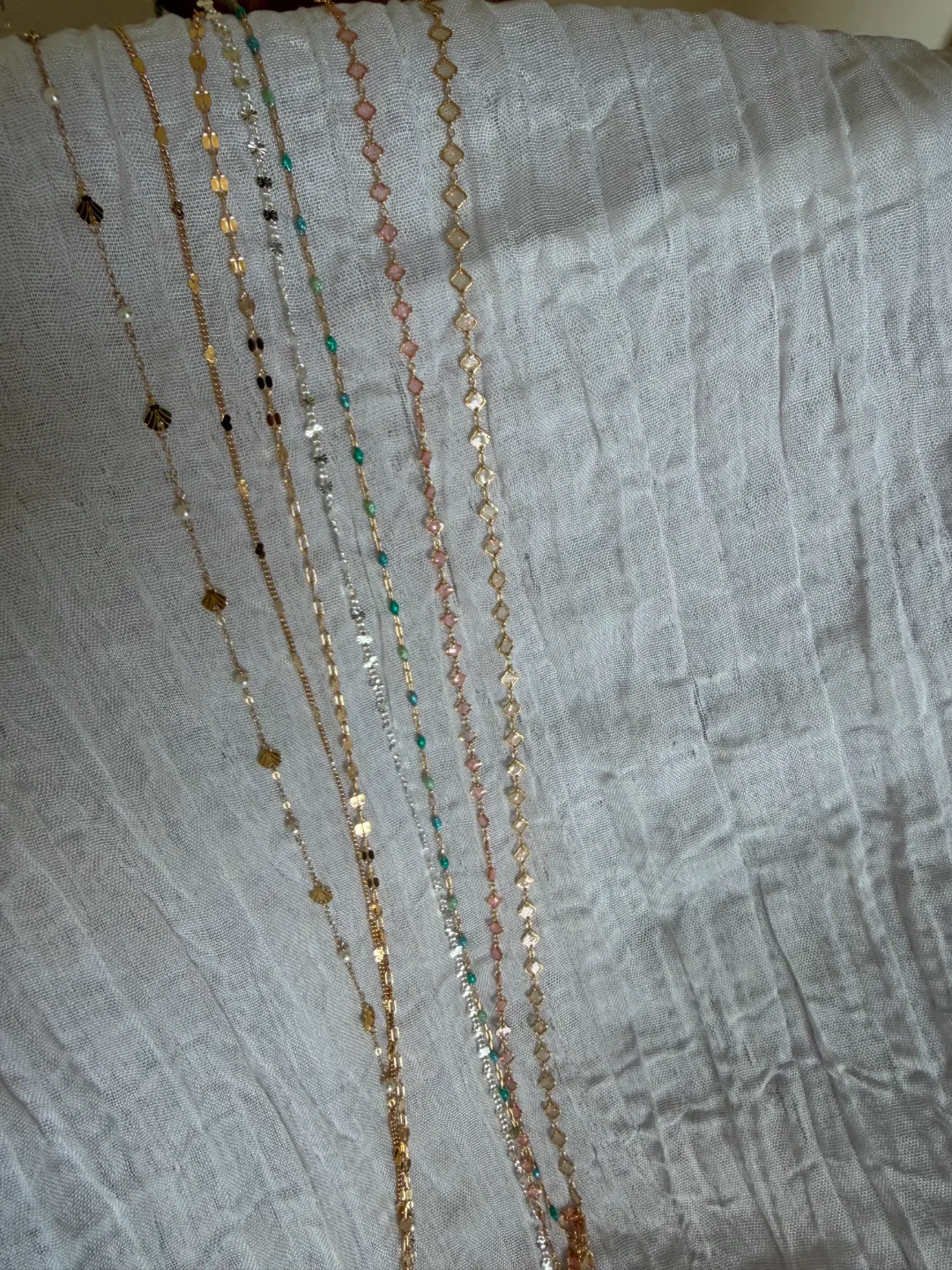 A lineup of gold and pastel-beaded permanent jewelry chains laid out on soft white linen