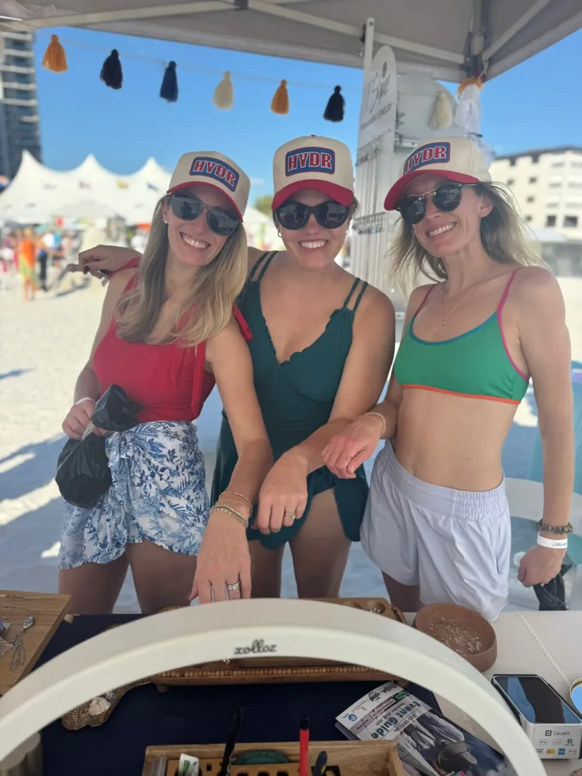Three friends smiling under a beach tent showing off their matching permanent bracelets