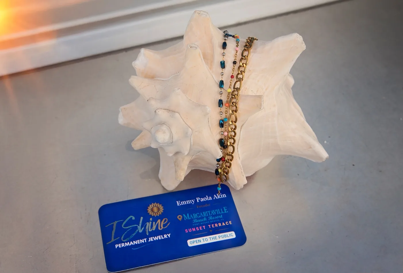 Blue and gold permanent chains draped over a conch shell beside an I Shine Margaritaville Sunset Terrace card